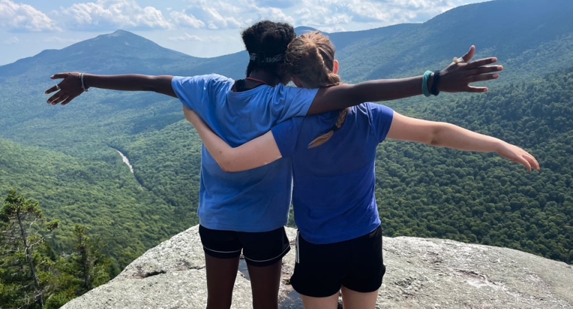 Two students standing on a rocky overlook face away from the camera toward the mountainous landscape below and wrap their arms around each other. 